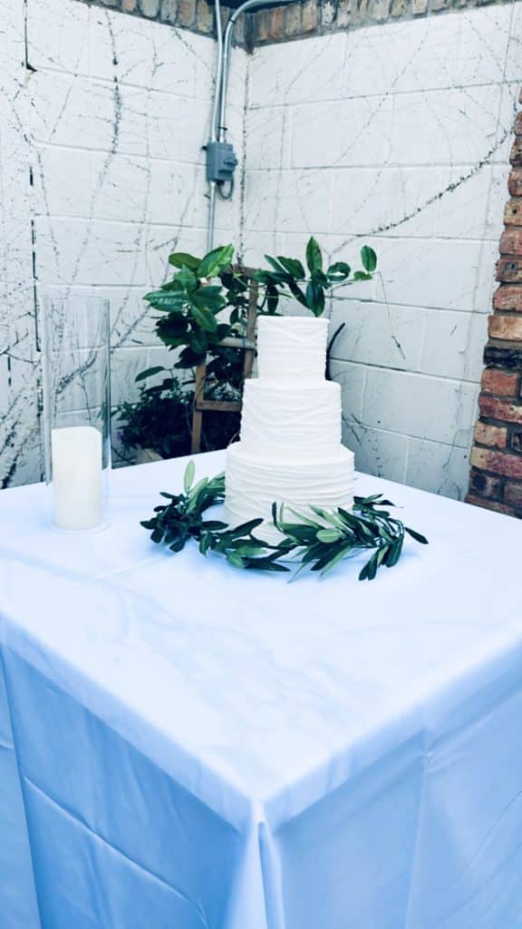 three tiered white cake, simply adorned with lemon leaves around the base on a white tablecloth and a single candle in a glass vase with a loom tree behind it, Central FL