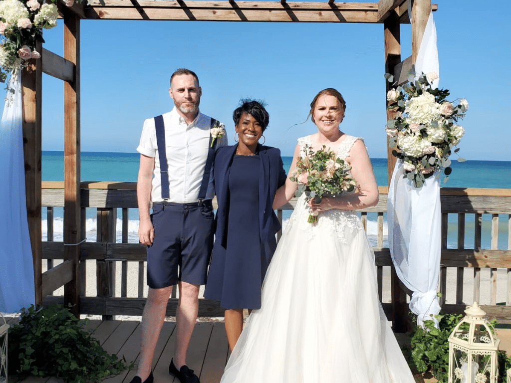 Bride and Groom with their celebrant outdoors under a pergola, wrapped with white fabric, outdoors, officiant in a blue dress suit, groom wearing dark shorts and suspenders, bride in a long flowing white wedding dress, Central FL