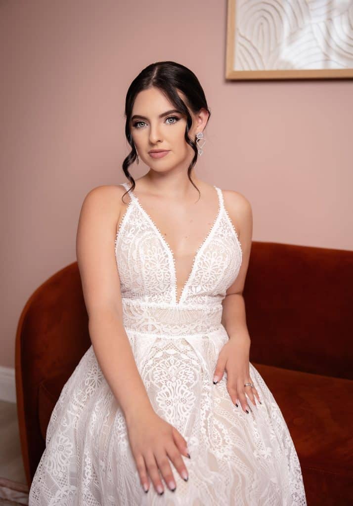 Burnette bride wearing a white lace dress, sitting on a red sofa, up do with soft make up, curls shaping her face, Haley Finegan Hair and Make Up, Central FL