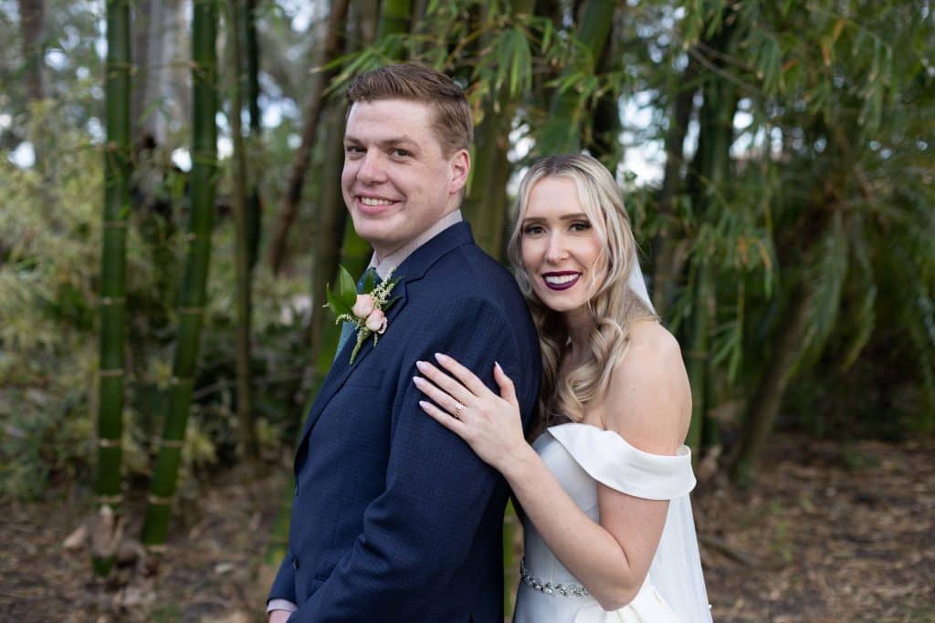 Bride standing behind her groom in the woods, her left hand on his shoulder, bride in her white gown, groom in a blue suit, Central FL