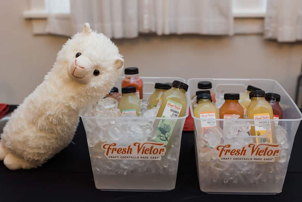 two plastic tubs sit on black table with ice and bottles of juice on top of it with plush llama leaning against them