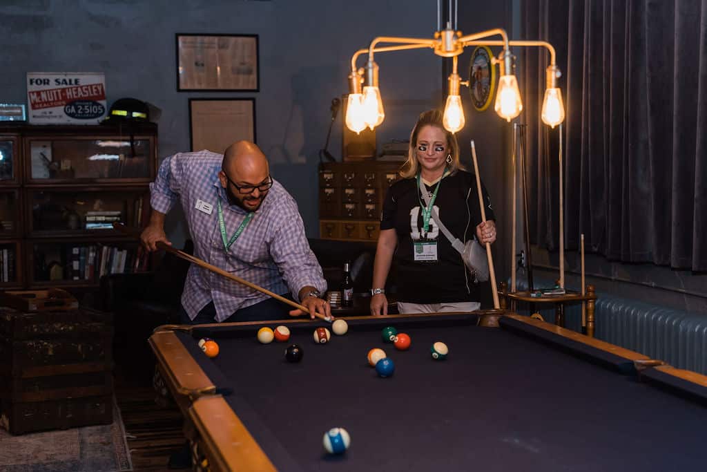 man lines up shot at pool table with woman holding a pool stick stands beside him watching