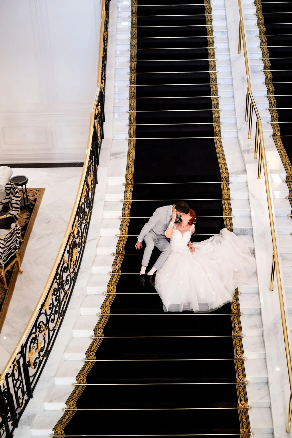 Bride and groom sharing a romantic kiss while seated on the grand marble staircase with black and gold carpet runner at World Equestrian Center in Ocala, Florida, the bride wearing a full ballgown with lace bodice and cathedral train as her groom in a gray suit leans in for a kiss photographed from above