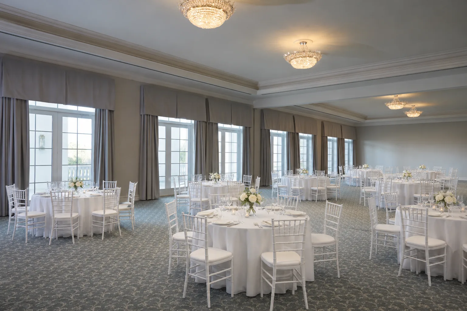 Elegant ballroom reception setup at World Equestrian Center in Ocala Florida featuring white chiavari chairs, round tables with white linens, white floral centerpieces, crystal chandeliers, and floor-to-ceiling French doors with neutral drapery