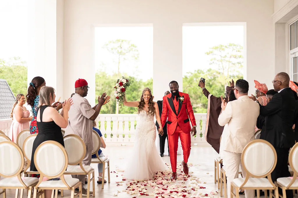 Bride and groom walking down the aisle after their ceremony at World Equestrian Center in Ocala Florida, groom wearing a bold red suit and bride in a lace ballgown holding a red and white floral bouquet as guests celebrate along the rose-petal-lined aisle