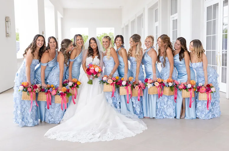 Bride in white lace gown surrounded by eleven bridesmaids in light blue satin and floral applique dresses holding colorful floral arrangements and wicker baskets with pink ribbons at World Equestrian Center in Ocala Florida