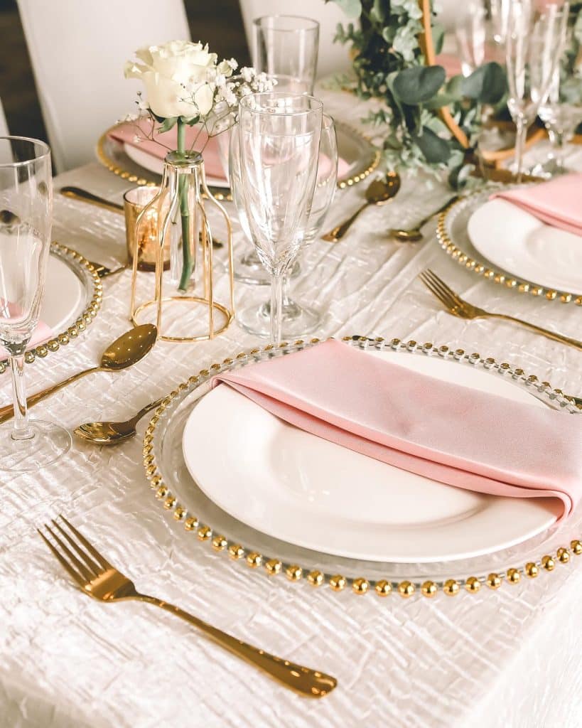 tables cape with white tablecloth, textured off white runner, clear chargers with gold beaded borders, white plates, with pink napkins and gold flatware, accompanied by clear glassware and a floral centerpiece, Creations Cuisine Inc, Central FL