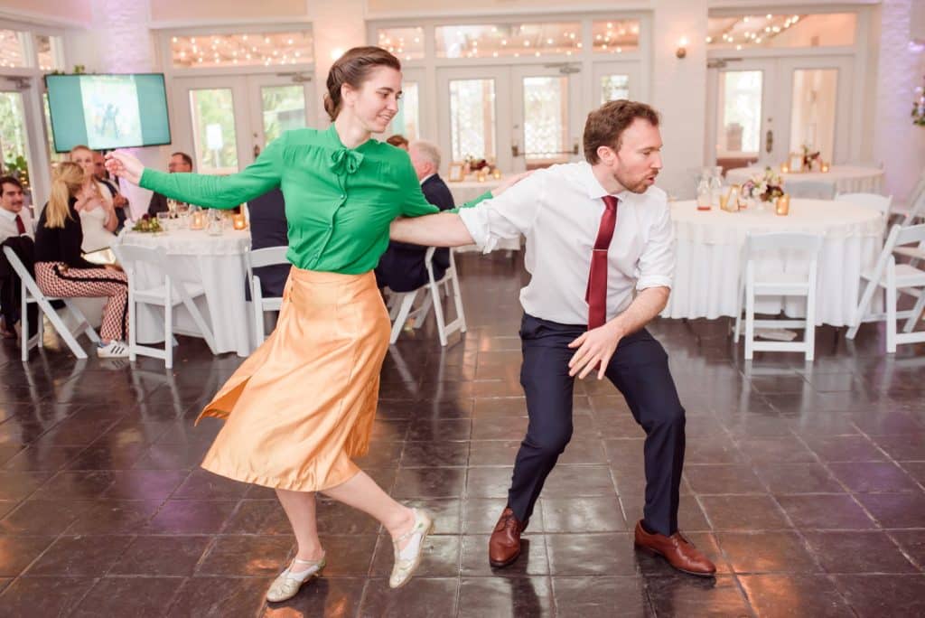 Couple dancing indoors in a room with windows in the back with tables and chairs behind the couple, woman wearing a green blouse, gold skirt, man wearing blue pants, white shirt with a red tie, Vintage Dance Company, Central FL