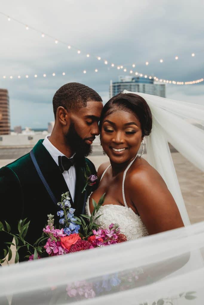 Black couple in their wedding attire, outside on the beach, groom stealing a moment with this bride as she smiles, string lights in the backdrop, skyscraper in the distance, Central FL