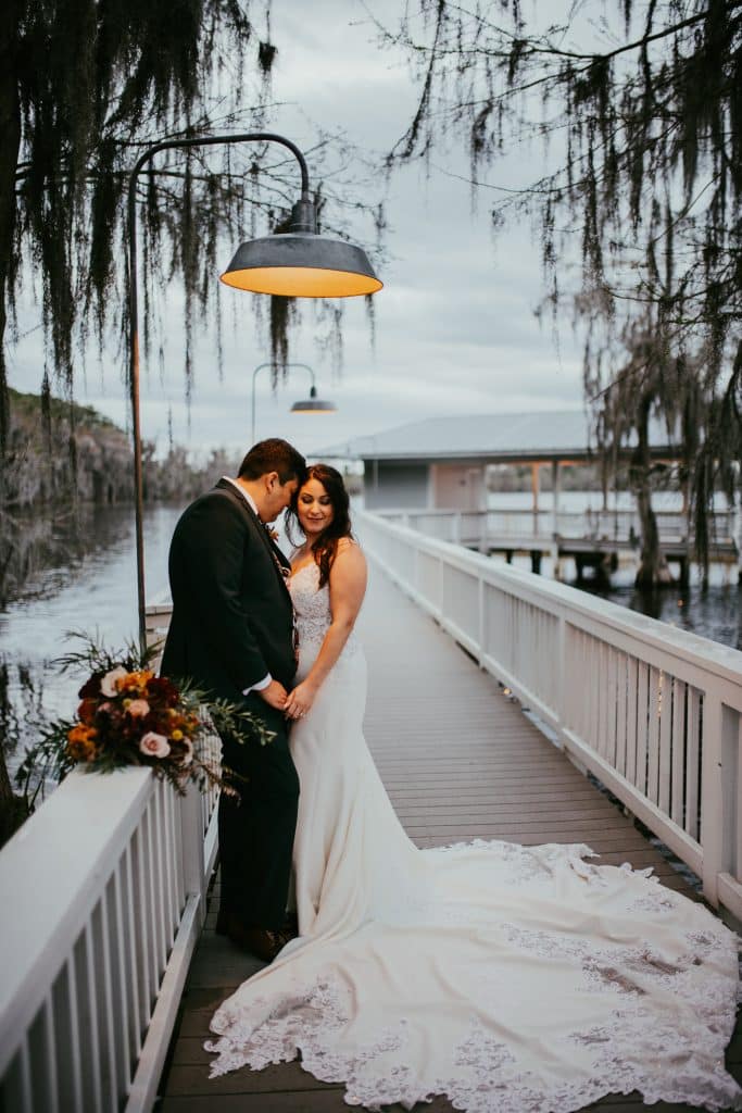 Wedding couple on a bridge near the water, under a lamp, he is looking down at her while she looks towards the camera, taken at dusk, Central FL