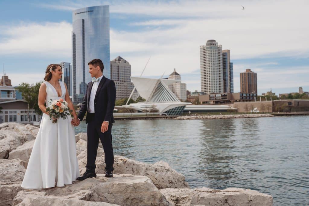 Wedding couple facing each other and standing at the water's edge on rocks, with the city in the background, Ashley Krug Photography, Central FL