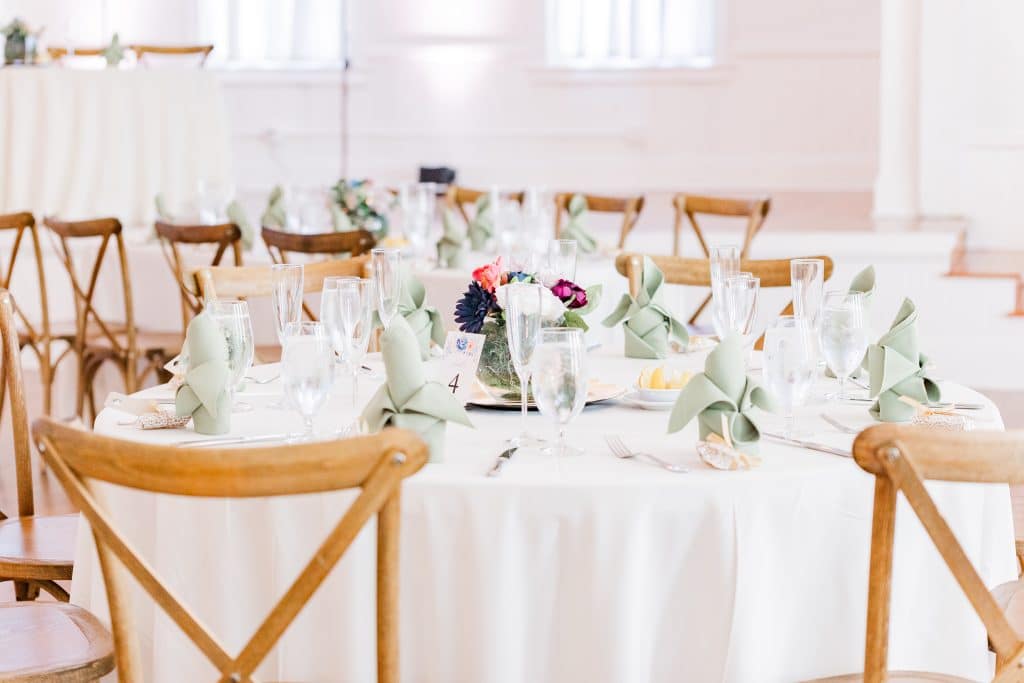 Tabelscape of a reception, white tablecloth, brown wooden chairs, mint green napkins, clear glassware, vibrant colored floral arrangement as a centerpiece, Central FL
