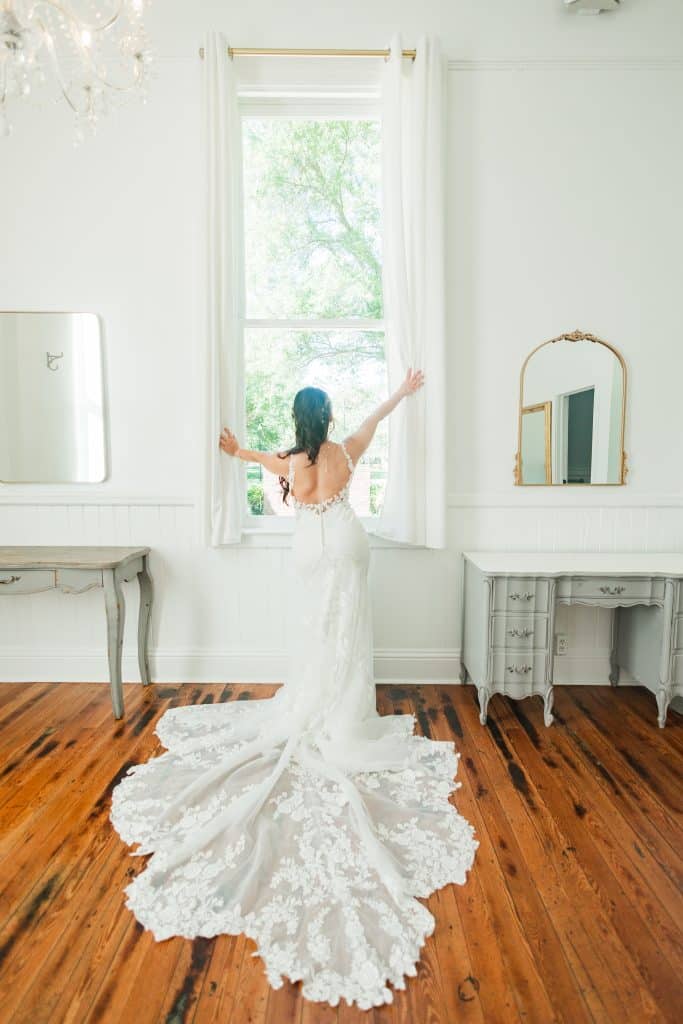 A bride taking a peek out the tall window in her wedding dress, with her train spread out on the floor to show the intricate lace design, Central FL