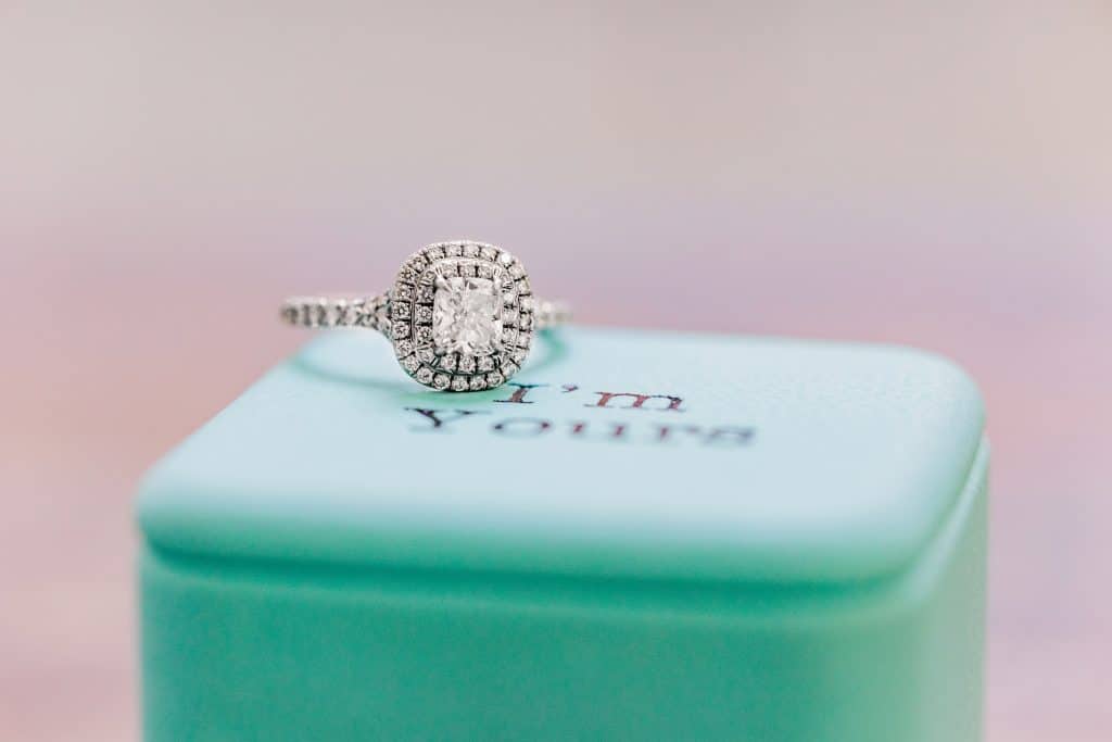 Flatlay of an engagement ring sitting atop a teal box that says 