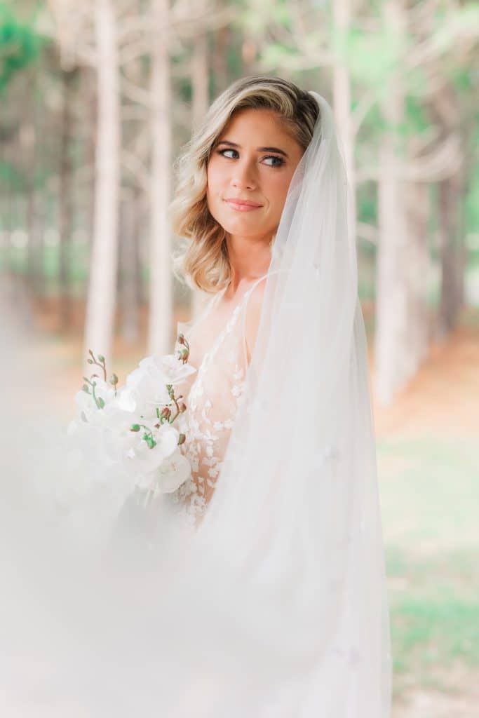 Bride looking over the shoulder of the photgrapher, with her veil in the breeze to partially blur the photo, outdoors in a wooded area with trees in the background, Central FL