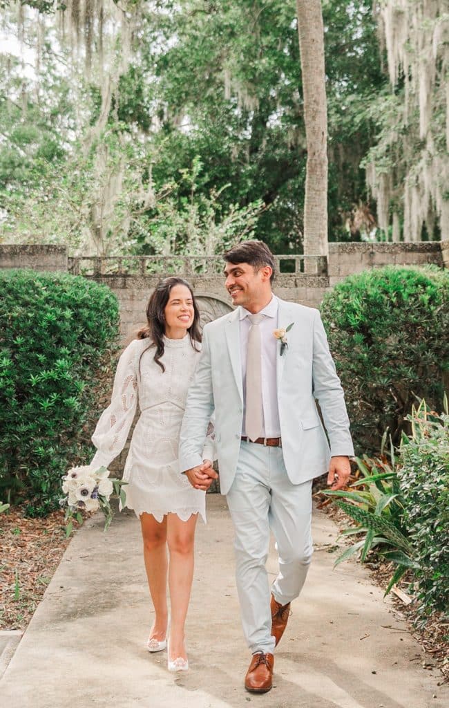 Engagement photos shoot, couple walking towards the camera, in a garden, woman in a short white laced dress, man in a light grey suit, Central FL