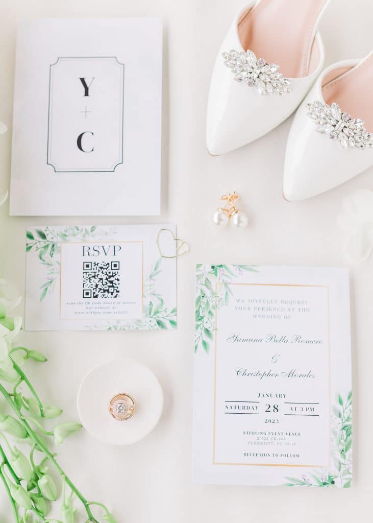 Flatlay of wedding invitations, RSVP card, pearl earrings, wedding shoes with silver embellishments, a ring tray displaying the rings and stationary with the couple's initials on them, Central FL