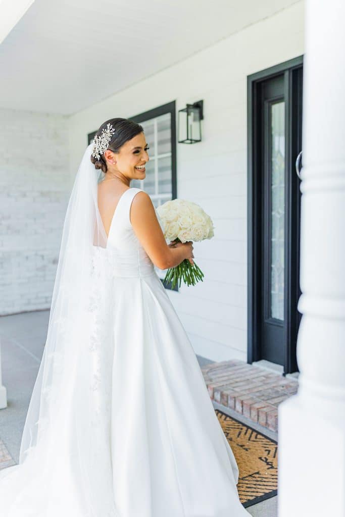 Beautiful bride showing off her dress right before the ceremony, long white princess style gown, with a silver hairpiece to hold her white veil, standing outside a front door of a home, looking over her shoulder and smiling, Central FL