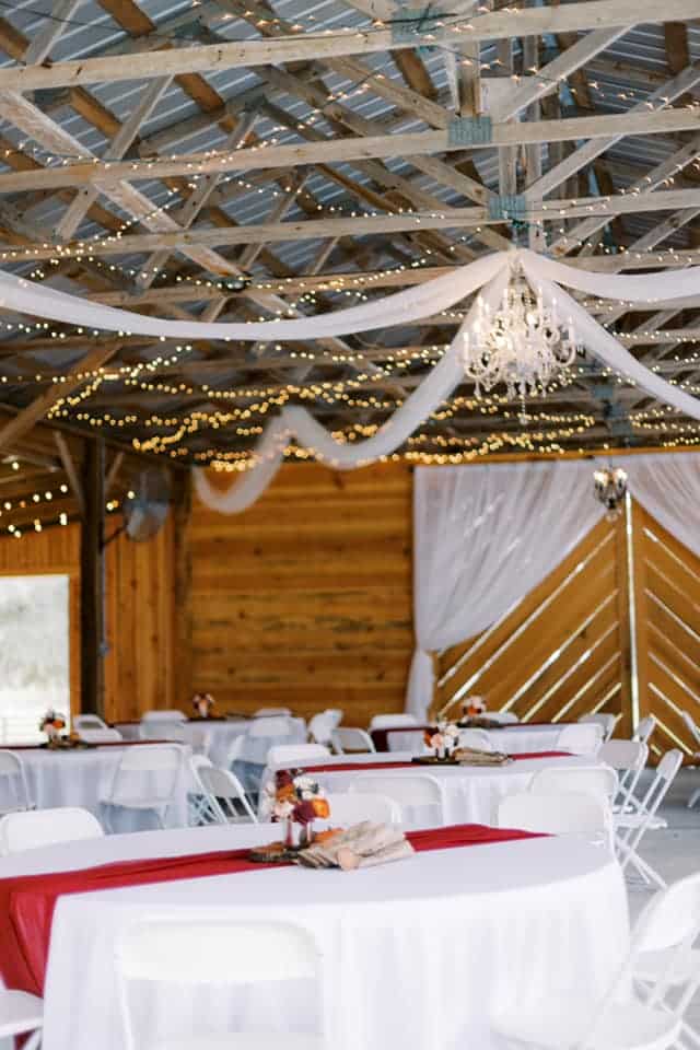 Indoor shot in a barn, wedding reception tables, white tablecloths, red runners, wooden walls in the backdrop, rafters have lights and white taffeta hanging from them, Peacock Ranch Wedding Venue, Orlando, FL