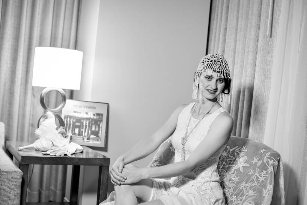 Black and White Photo, woman in a dress with a headpiece and long necklace, in a sitting room with a lamp on a table and seated in a chair, Vintage Dance Company, Central FL