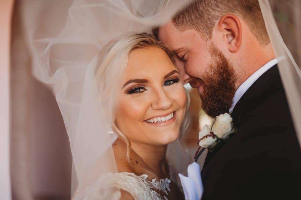 Close up of the bride with her groom, the groom whispering in her ear, while she smiles at the camera, her veil flowing above their heads, Ashley Krug Photography, Central FL