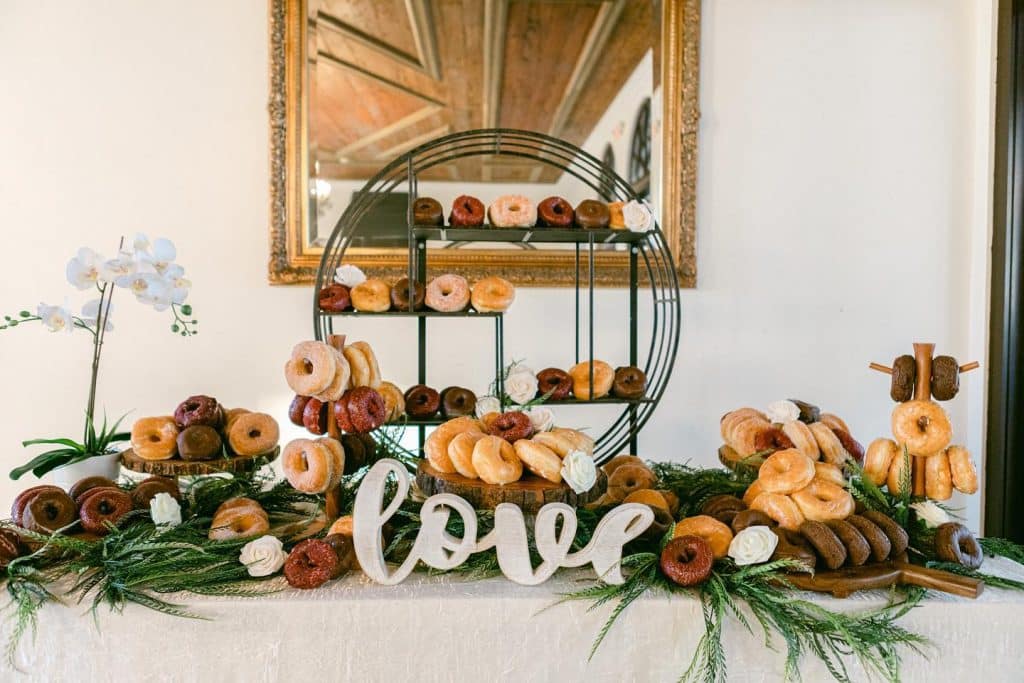Donut display with green stand and greenery around the donuts on the table, with a white tablecloth, the word love in white wood on the table in front of the food, Central FL