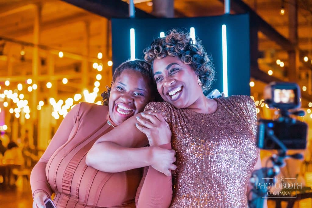 Two black women holding each other's arms, pose for the photo booth, one wearing a sparkling rose gold dress, the other wearing a mauve dress, The PhotoBooth Company, Central FL