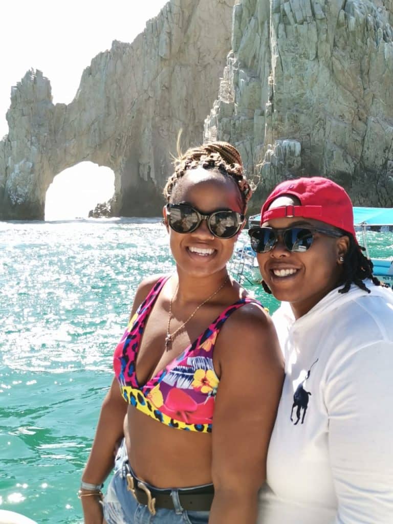 two young women sit together with sunglasses on in front of tropical body of water and rock formation behind them