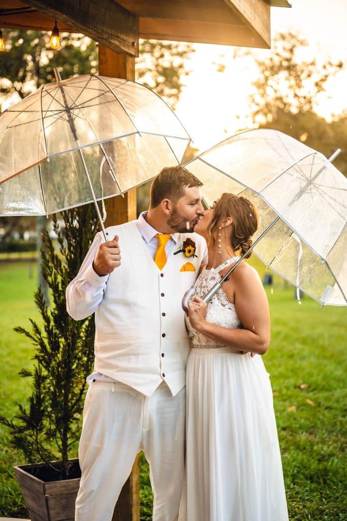 Wedding couple outdoors at sunset wearing all white, holding clear umbrellas while kissing, Orlando, FL