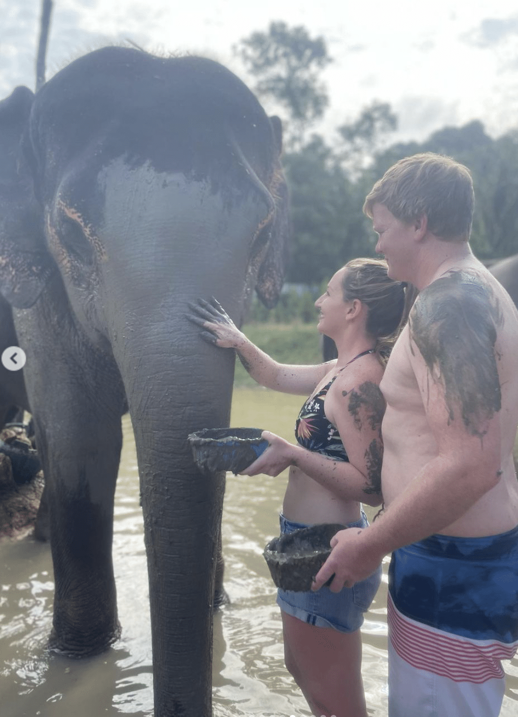 man and woman wear swimsuits while touching elephants trunk
