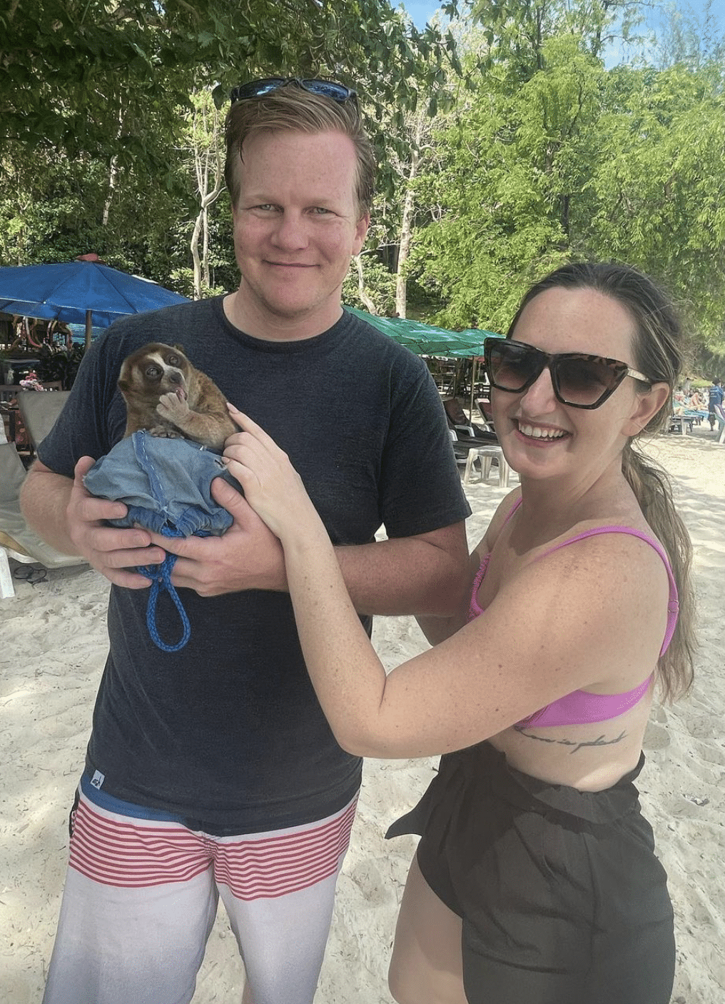 man and woman stand together on beach while man holds some sort of lemur