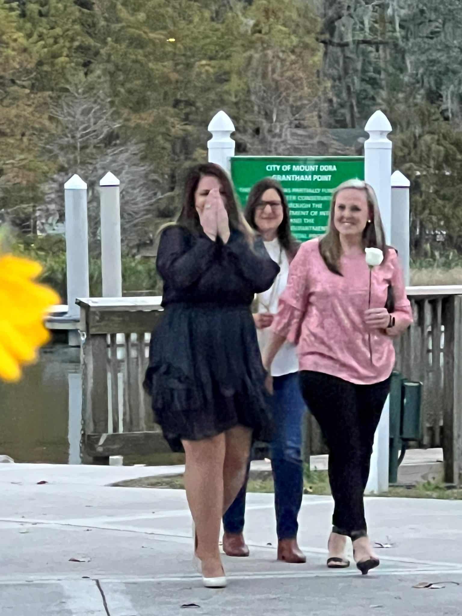 woman wearing black dress holds hands over mouth in surprise while two women walk next o her