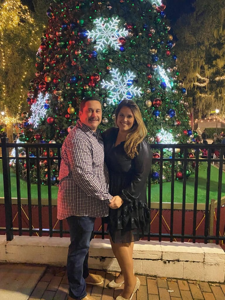 man and woman stand together holding hands facing each other in front of christmas tree with light up snowflakes decorating it