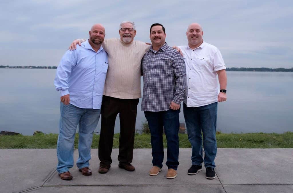 men in the family poses together smiling for picture in front of body of water