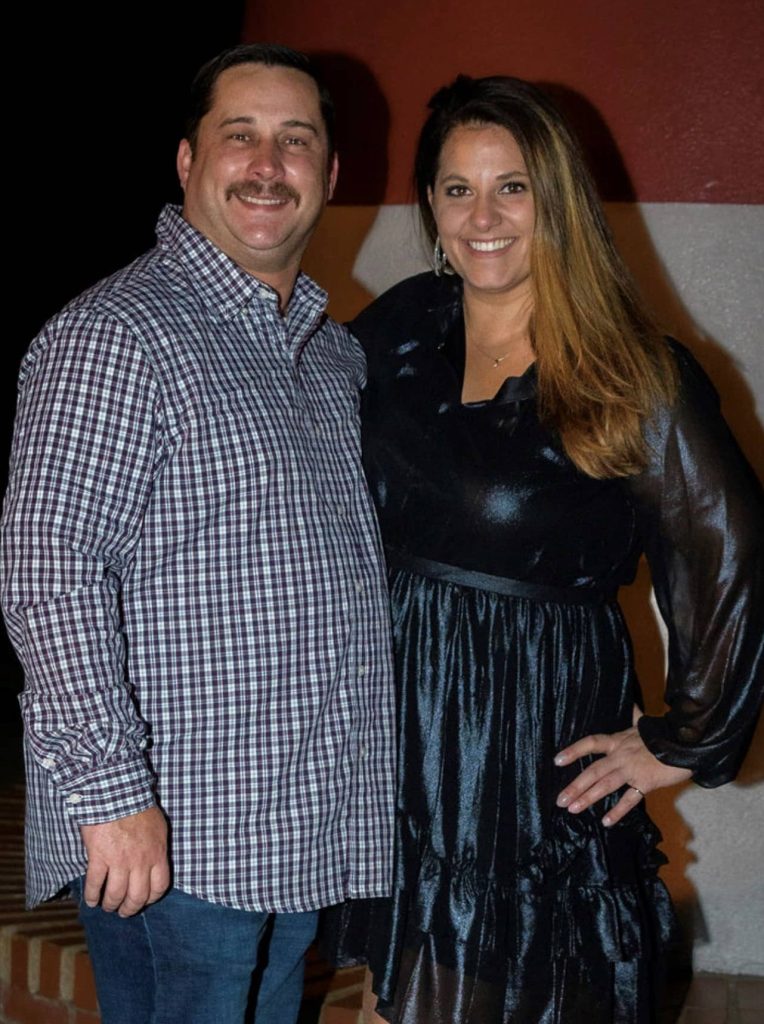 woman wearing black long sleeve dress stands next to man with mustache wearing button up long sleeve shirt