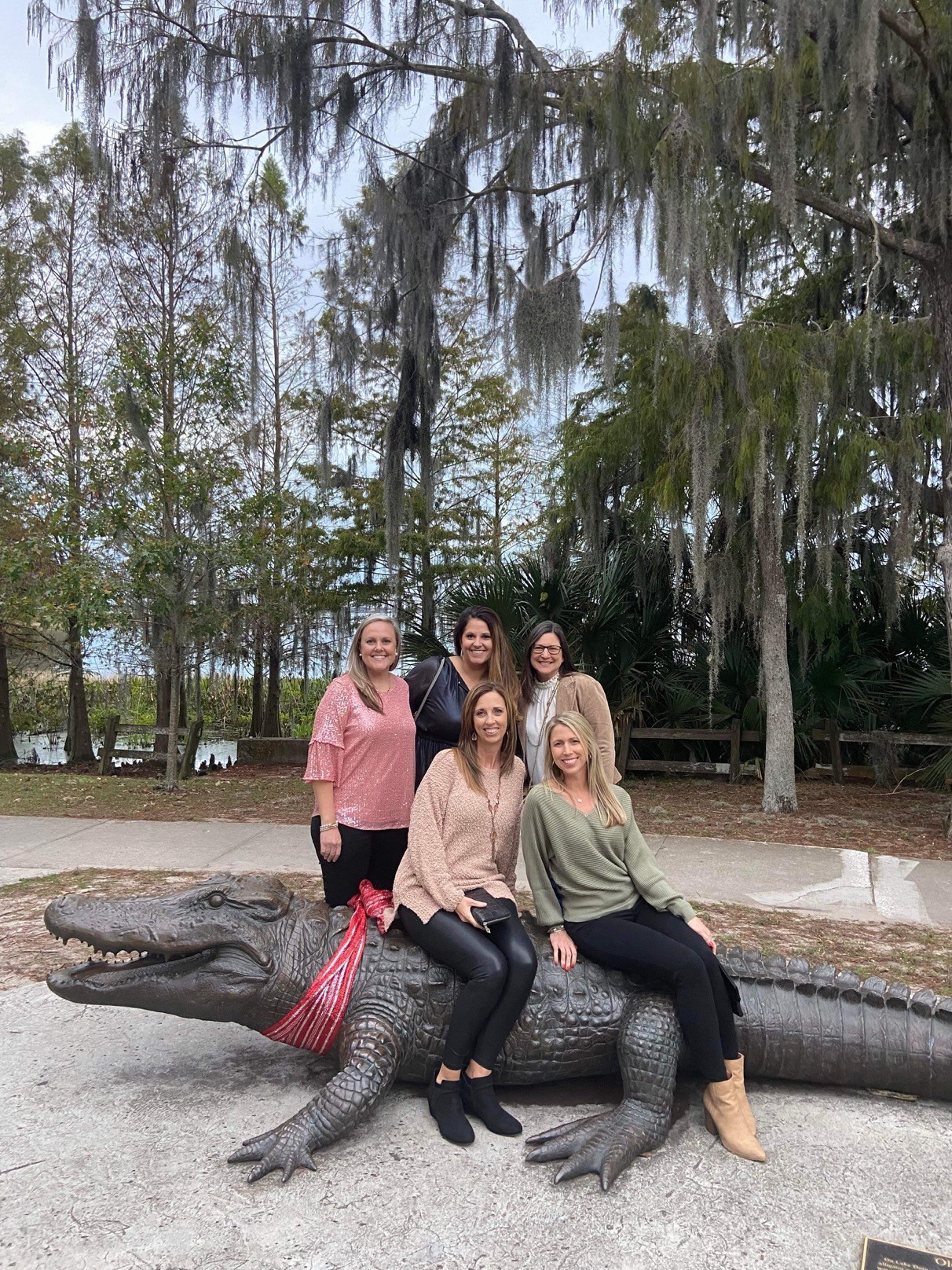 five women pose for picture some sitting on giant alligator statue and some standing behind them with trees in the background