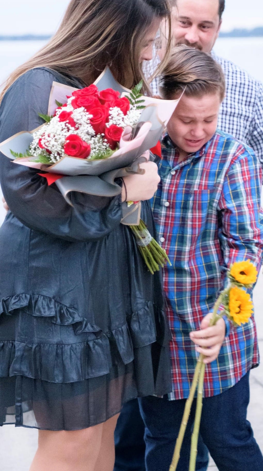 woman holds bouquet of white and red roses while hugging a boy in a plaid shirt holding sunflowers