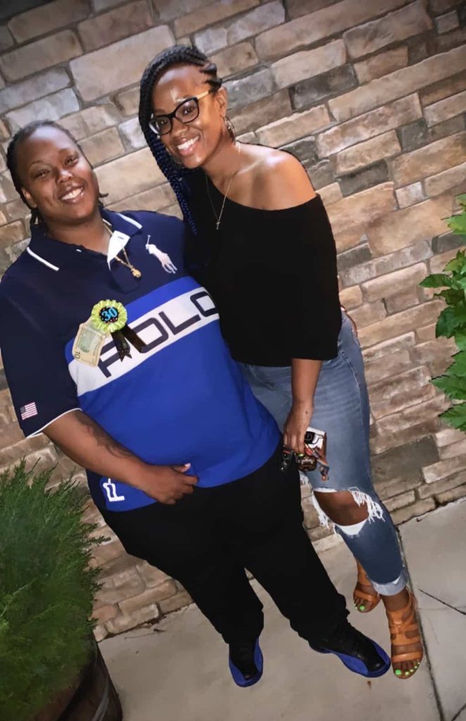 couple stands together smiling at the camera outside in front of brick wall