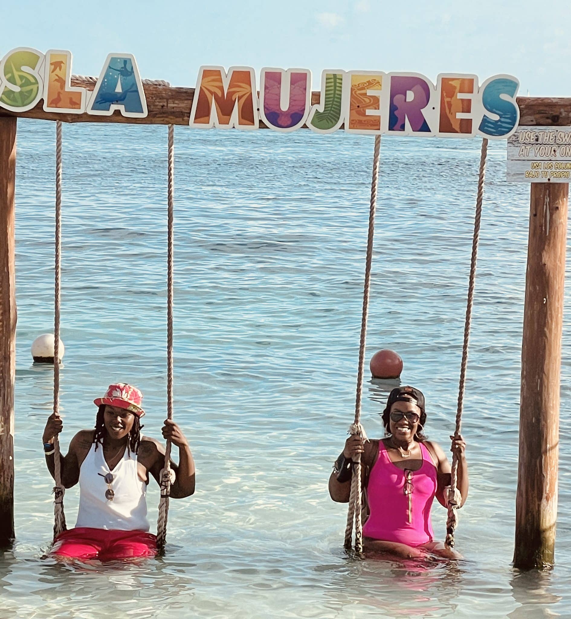 two women sit on swings in the ocean water with letters above reading isla mujeres