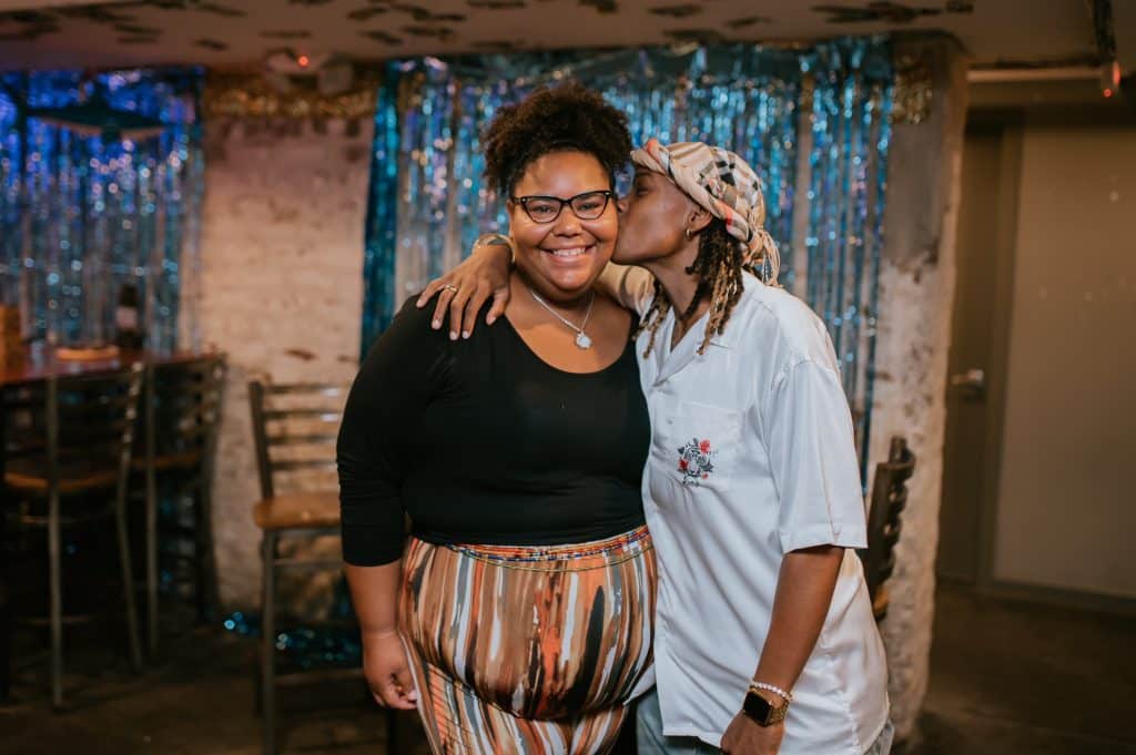 one woman stands smiling at the camera while the other leans over and kisses her on the cheek
