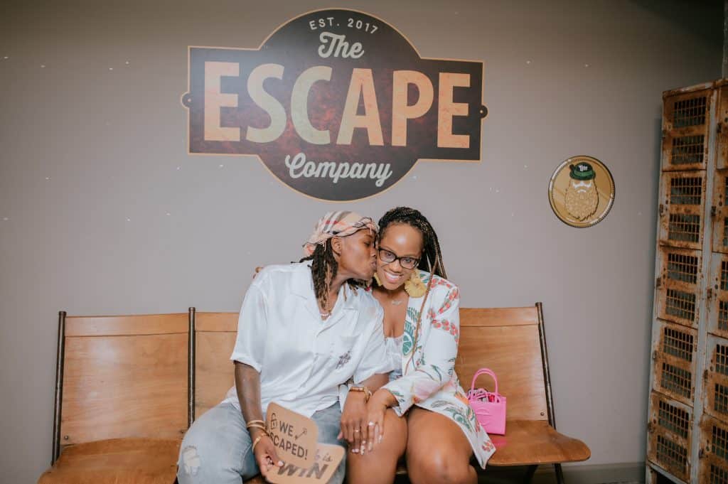 two women sit on the bench under a sign reading the escape company