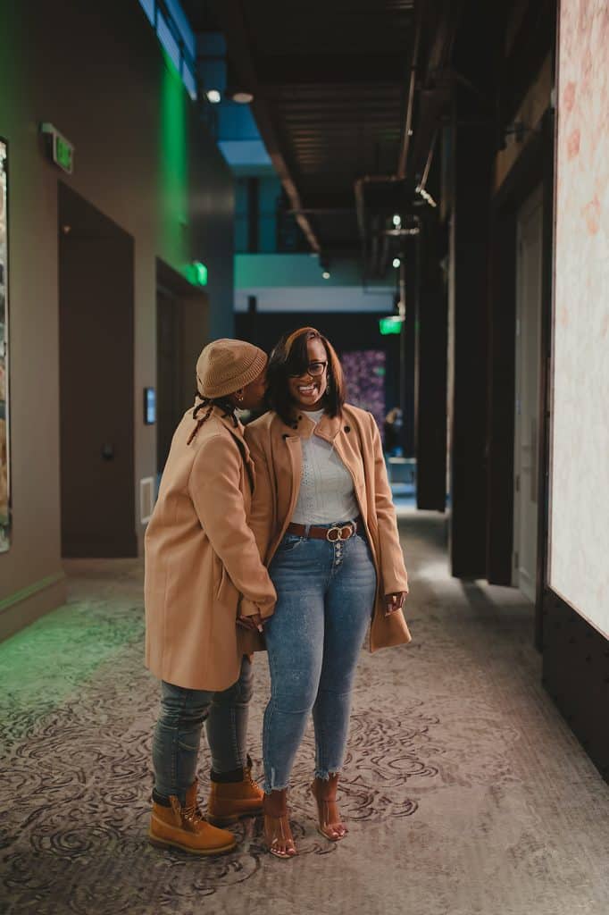 couple pose in matching outfits while one woman leans over to kiss the other womans cheek