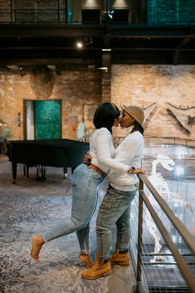 couple wearing matching blue jeans and white long sleeve shirts kiss while leaning against a railing inside