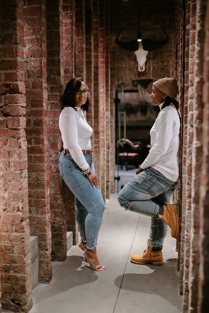couple wearing matching blue jeans and white long sleeve shirts post leaning against brick walls