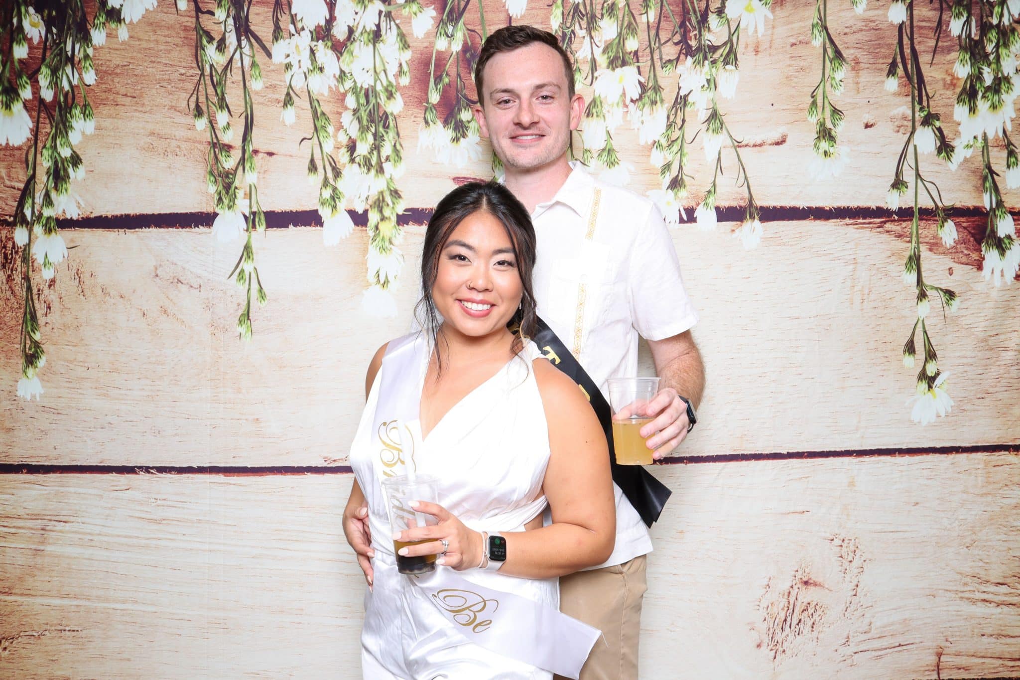 couple both wearing white stand together holding drinks in front of backdrop