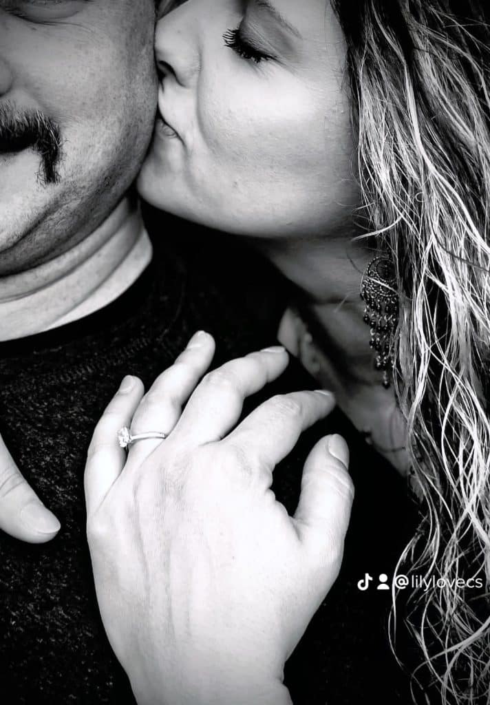 back and white image on woman kissing man on the cheek while her hand is on his shoulder showing off engagement ring