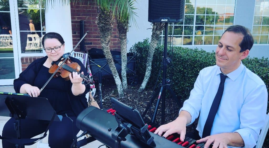 Two members of Rachel Durrum Music LLC, one on keyboard, one on violin, sitting in front of music stands, outdoors at an event, Orlando, FL