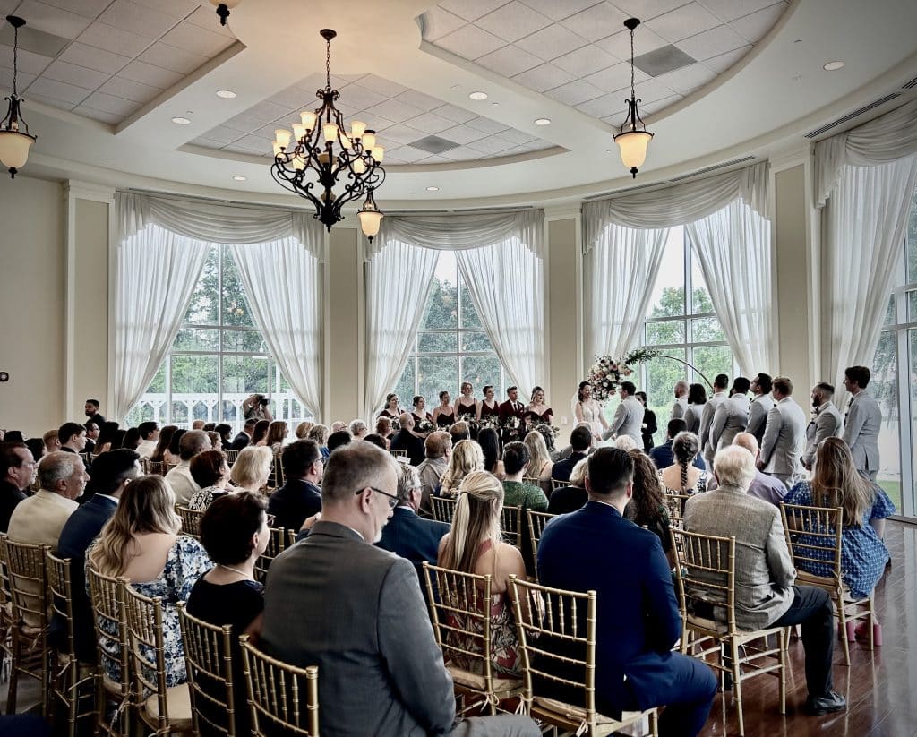 zoomed out shot of the wedding ceremony, bride, groom and wedding party at the front of the room, guests in seats in rows behind, large tall windows with white curtains, Rachel Durrum Music LLC, Orlando FL