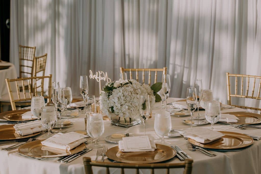 Tablescape for a reception, white tablecloth, gold chargers, white napkins, white flower centerpieces, gold backed chairs, Serenity Events, Orlando, FL
