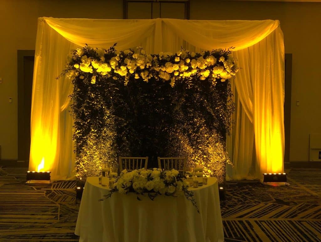 Head table set up with yellow uplighting and a white background behind the table, white flowers on the table with flowers and greenery on the white background, Dash of Class Platinum, Orlando, FL